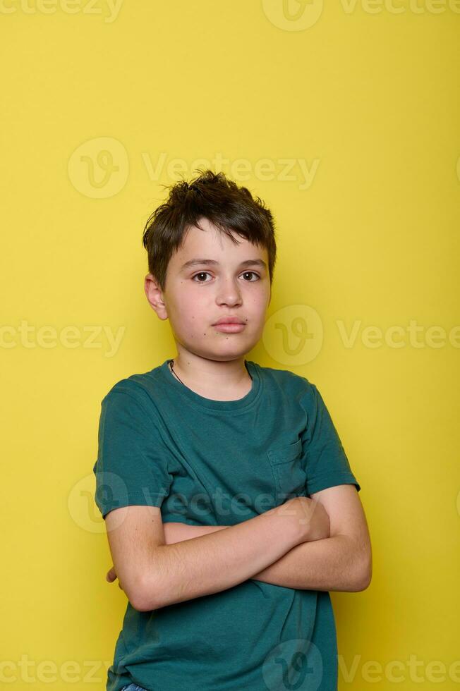 Emotional portrait of a sorrowful, overwhelmed school boy expressing sad emotions, with arms folded on yellow background photo