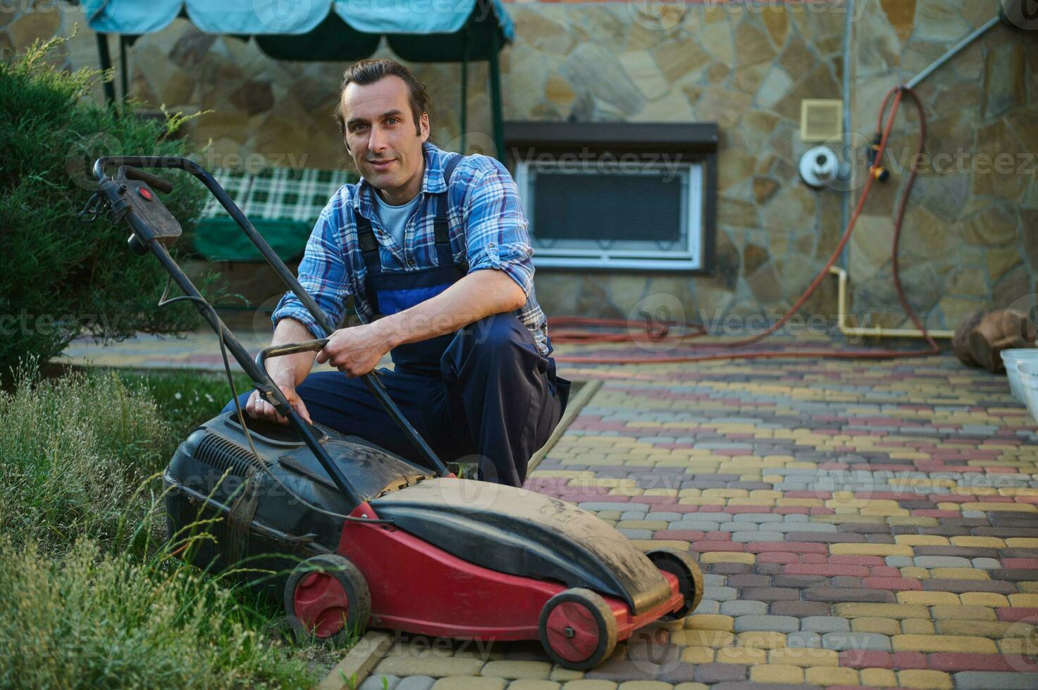 Attractive male gardener 40s, smiling looking at camera while replacing filter on electric lawn mower in the garden photo