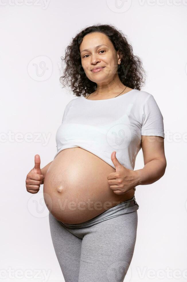 Multi ethnic beautiful pregnant woman gestures with thumbs up, expressing happiness and positive emotions photo