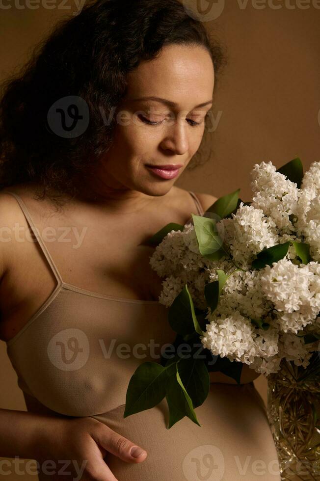 de cerca maravilloso grávido mujer en beige lencería, participación ramo de flores de floreciente lilas. el embarazo moda. cuerpo positividad foto