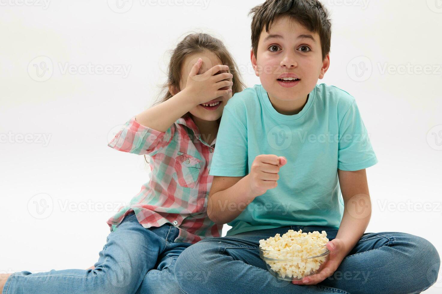 Amazed kids, teen boy and little child girl watching scary movie or cartoon, eating popcorn on white isolated background photo