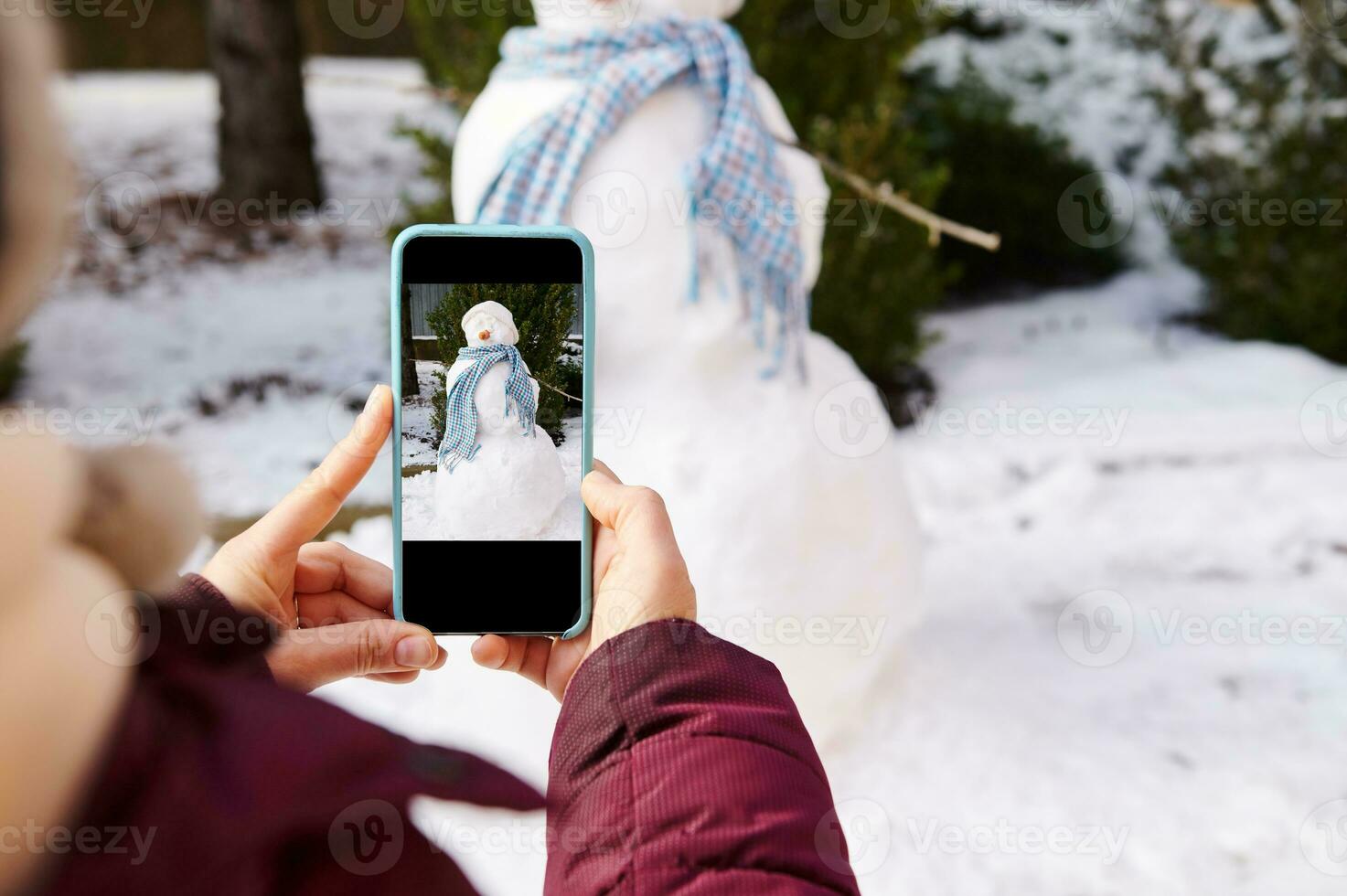 Closeup hands hold smartphone in live view mode, photographing snowman with blue shawl in snow covered nature background photo