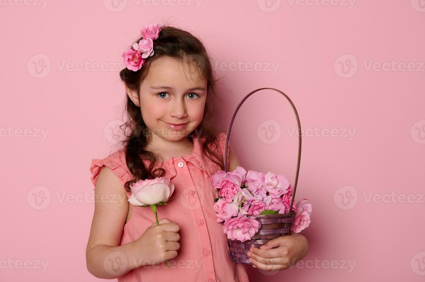 linda pequeño niña con Rosa flores en peinado, sonriente a cámara, posando con púrpura cesta lleno de Fresco rosado rosas, foto