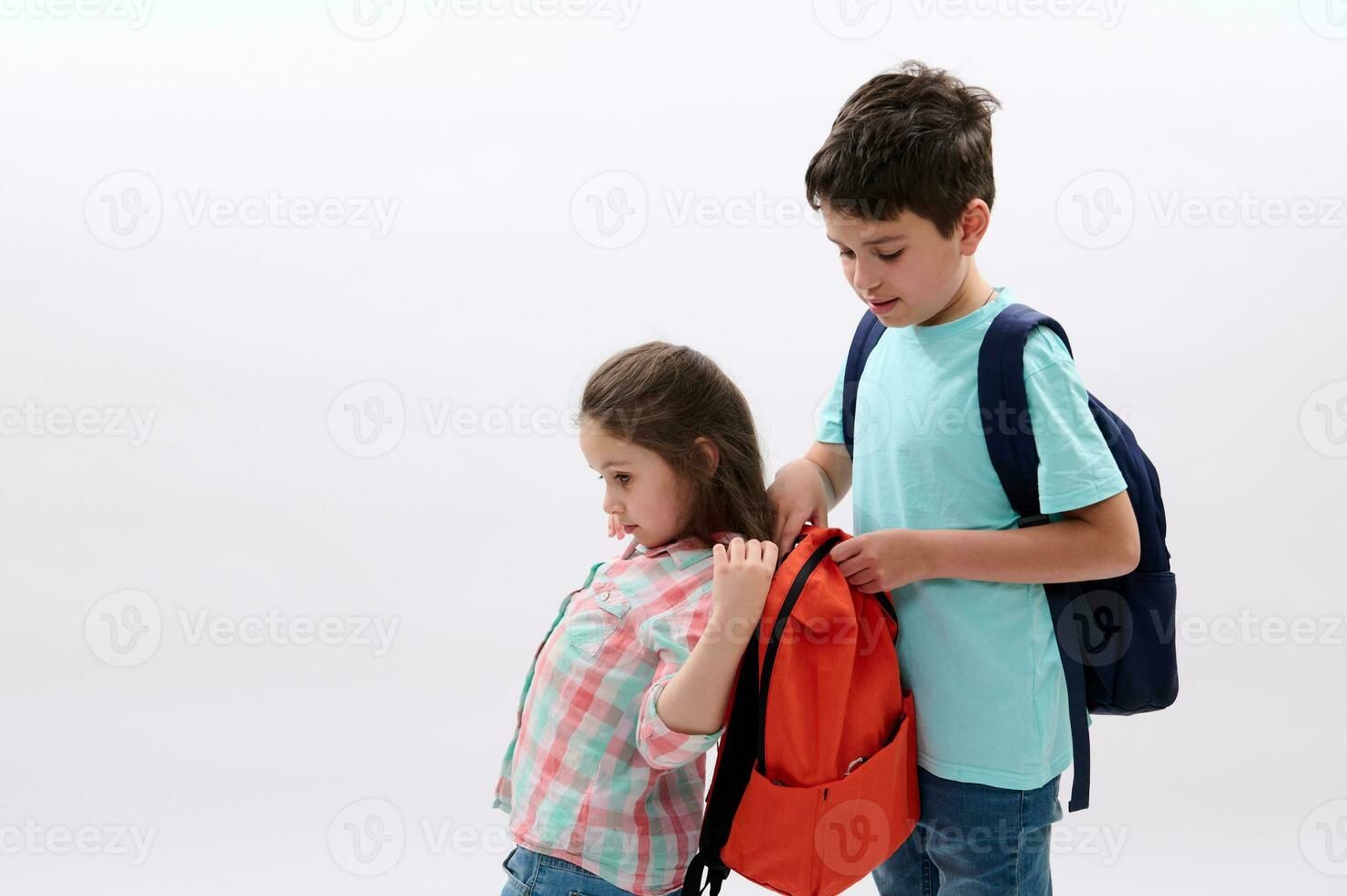 Caring teen boy older brother helps his younger sister to put on a backpack on back, isolated on white studio background photo