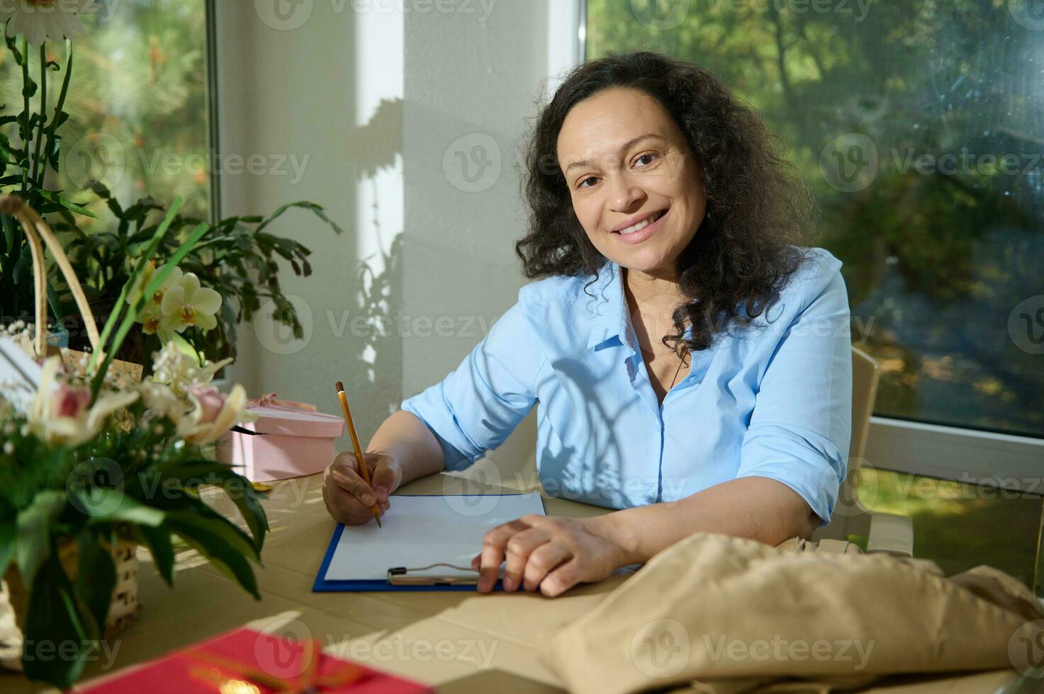 multiétnico encantador mujer trabajando en un floral comercio, sentado a mesa y escritura abajo notas, sonriente mirando a cámara foto