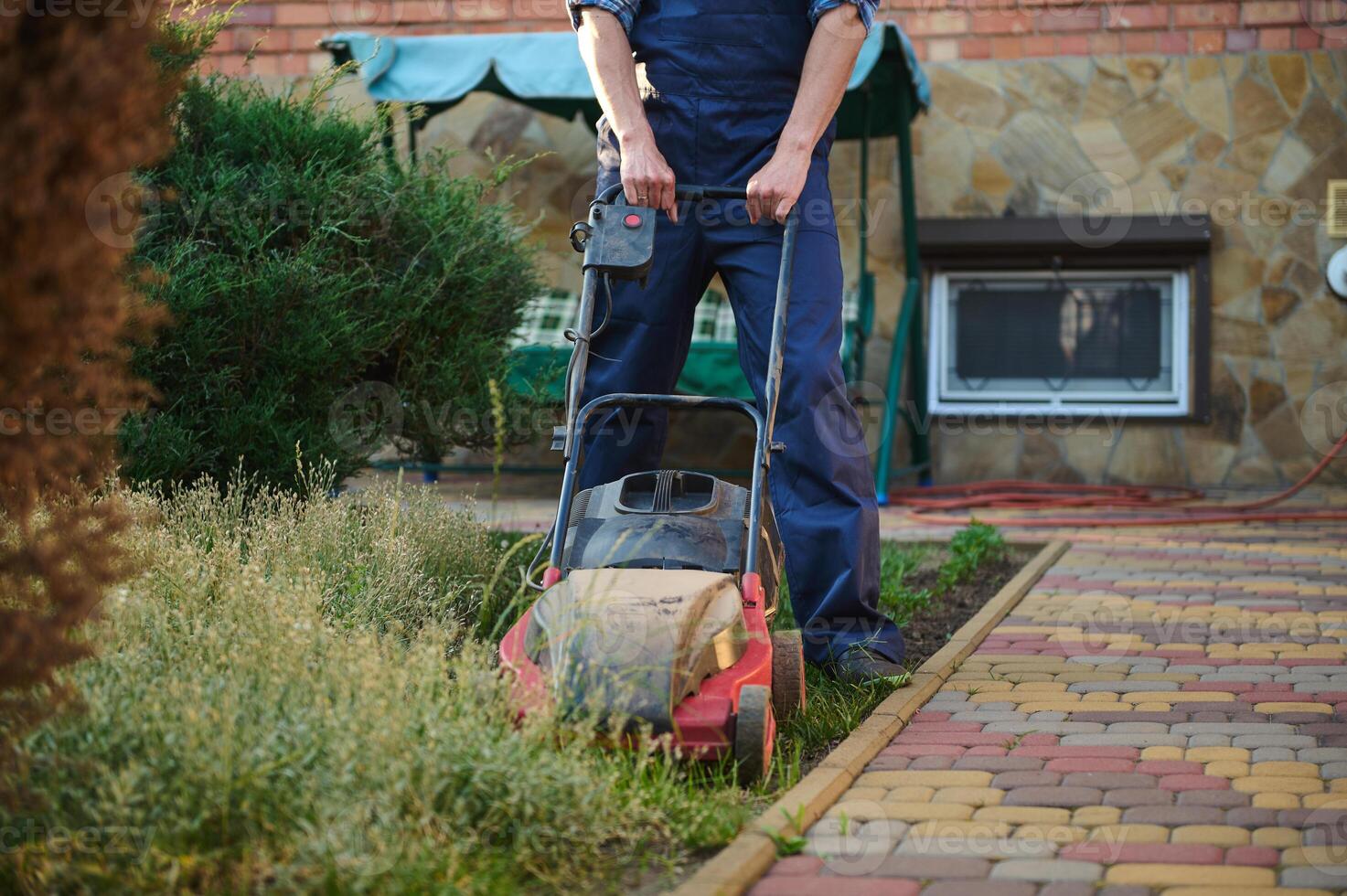 Close-up gardener in work uniform, mowing backyard grass, moving after a push electric lawn mower. Landscaping concept photo