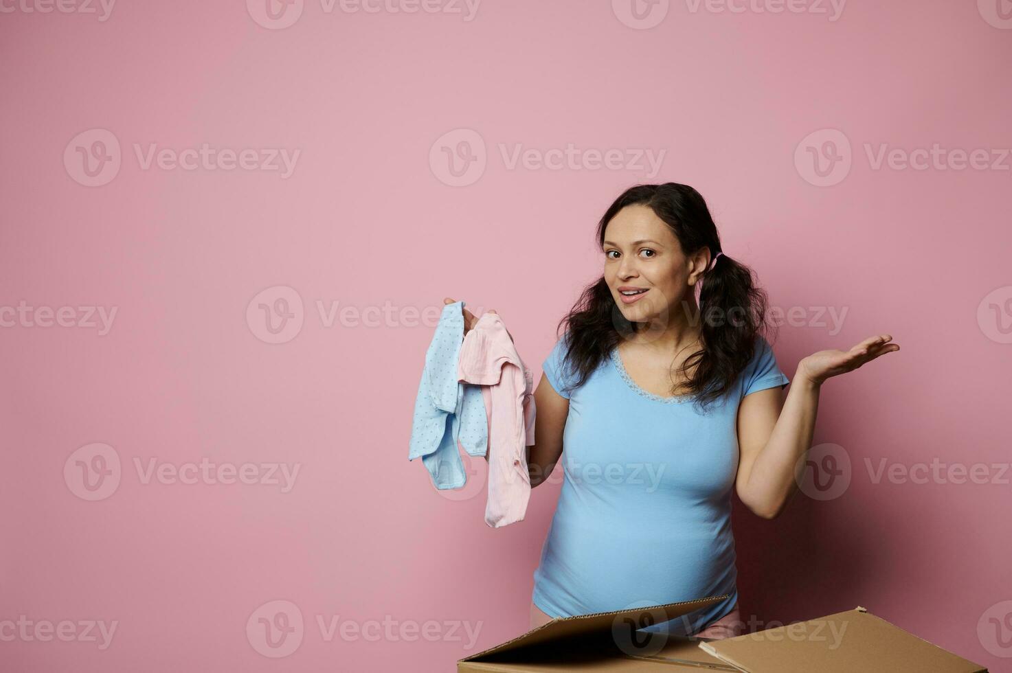 Happy pregnant woman with big belly shows blue pink newborn bodysuit. Gender reveal party, baby shower, isolated on pink photo