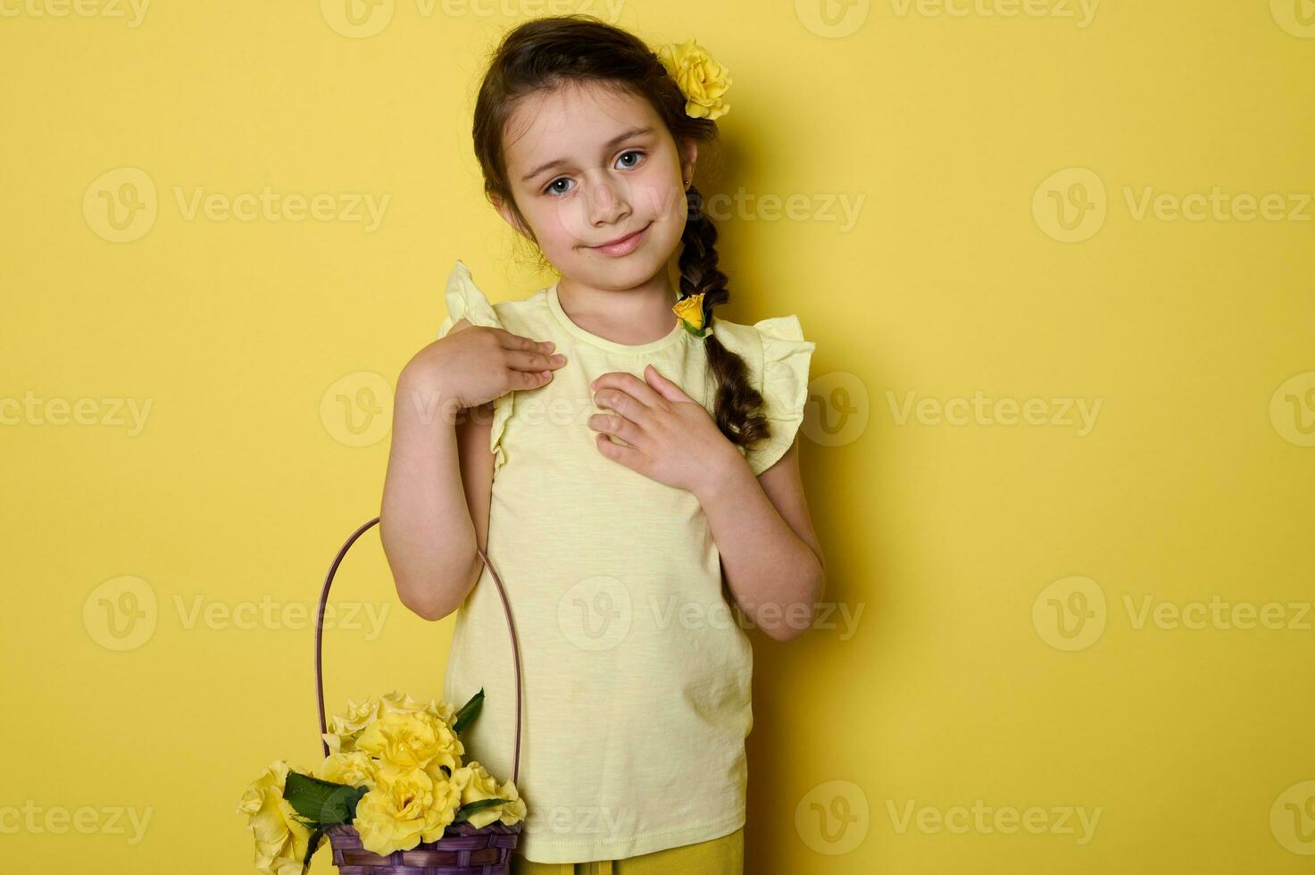 emocional retrato de adorable pequeño niño niña vistiendo amarillo vestido, participación púrpura mimbre cesta con amarillo rosas foto