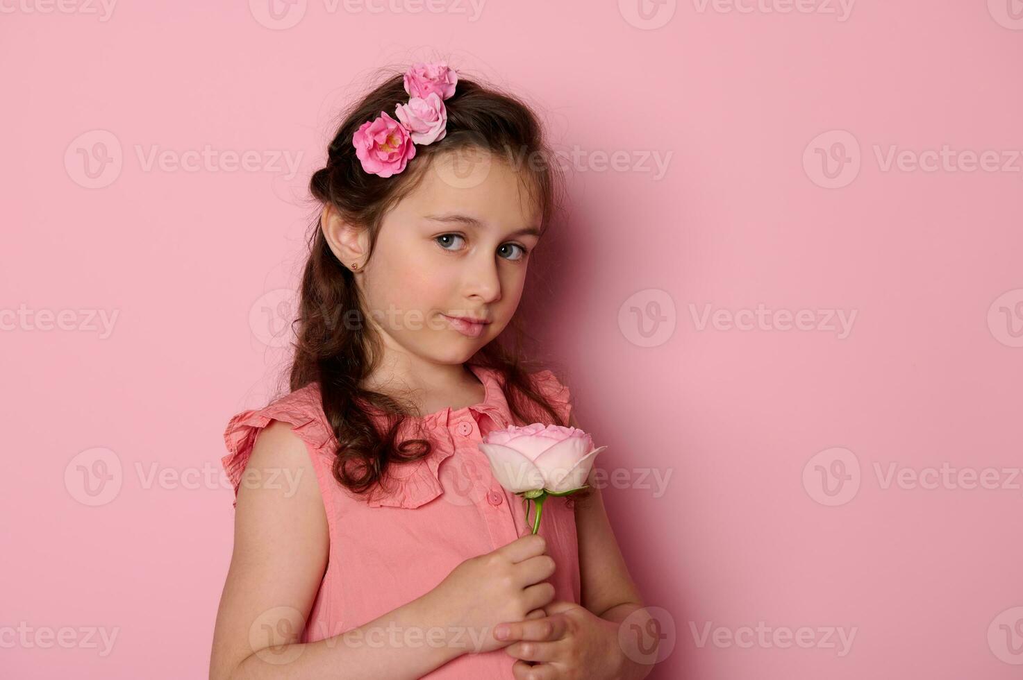 auténtico elegante pequeño niña en rosado vestido, participación un Rosa flor, sonrisas lindo mirando a cámara, aislado antecedentes foto