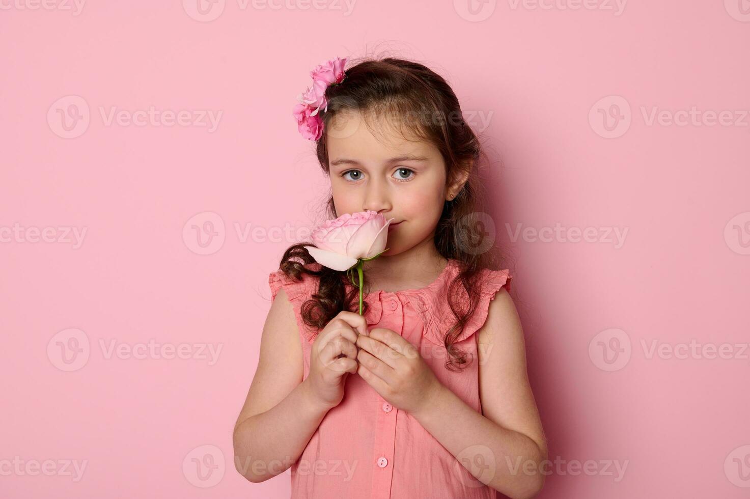 Portrait of a noble charming baby girl in pink dress and flowers in hairstyle, enjoying the smell of pink rose flower photo
