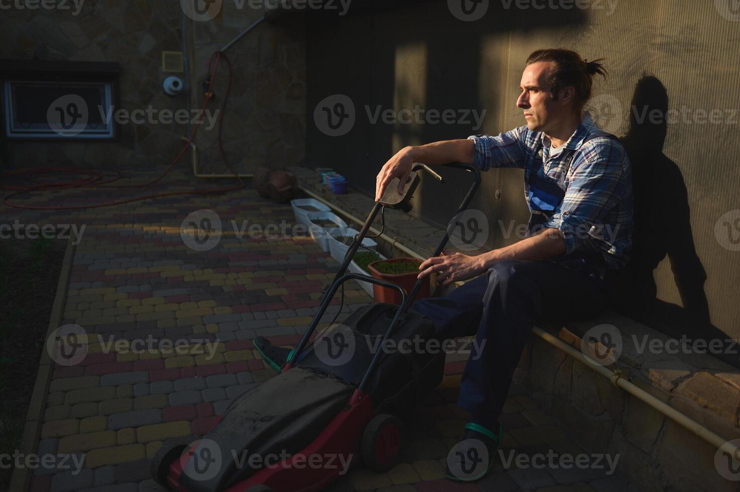 Portrait of a tired overworked gardener man with electric lawn mower, relaxing after hard working day in the backyard photo