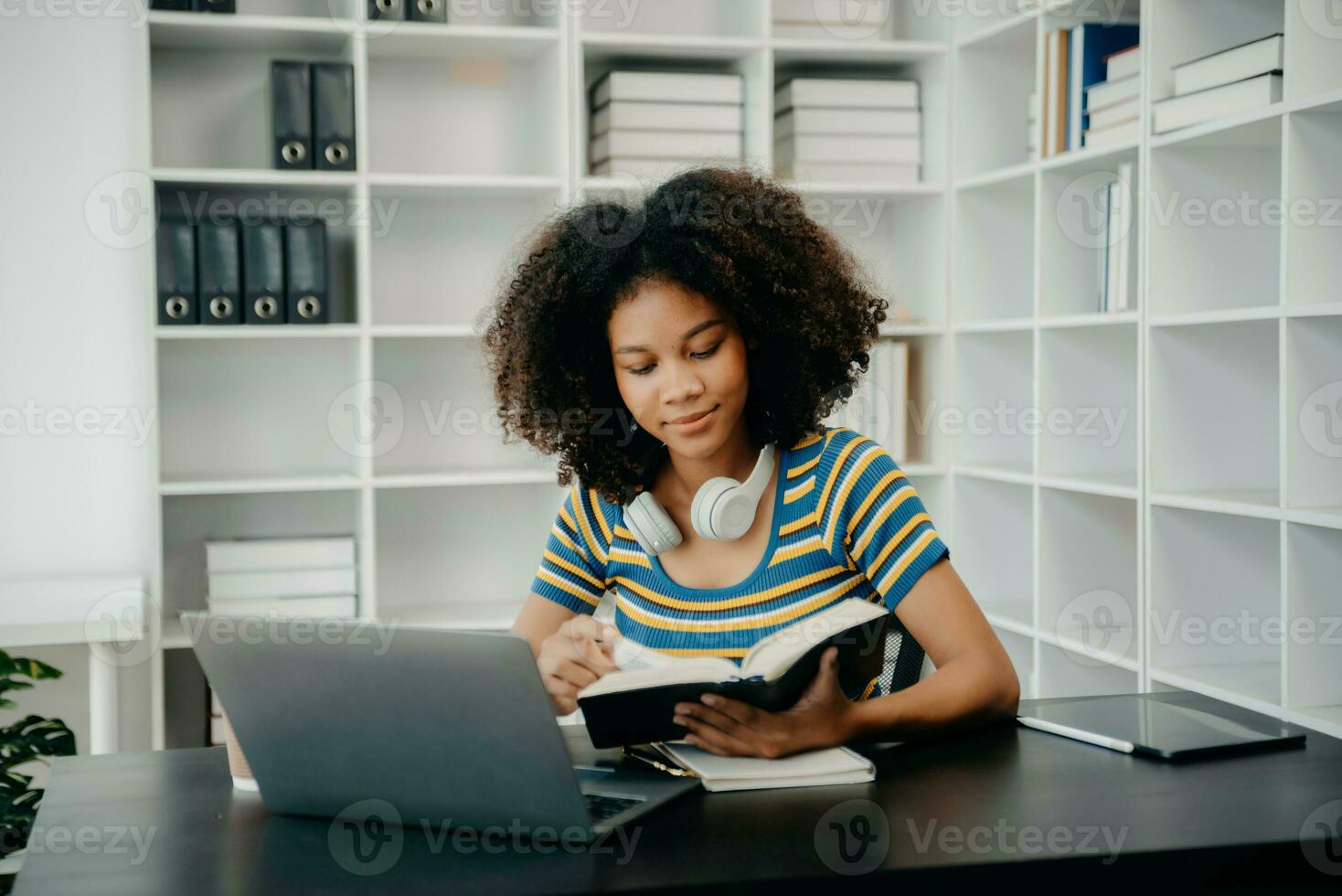 young African student studying at the college library, sitting at the desk, using a laptop computer, tablet and headphones having a video chat. photo