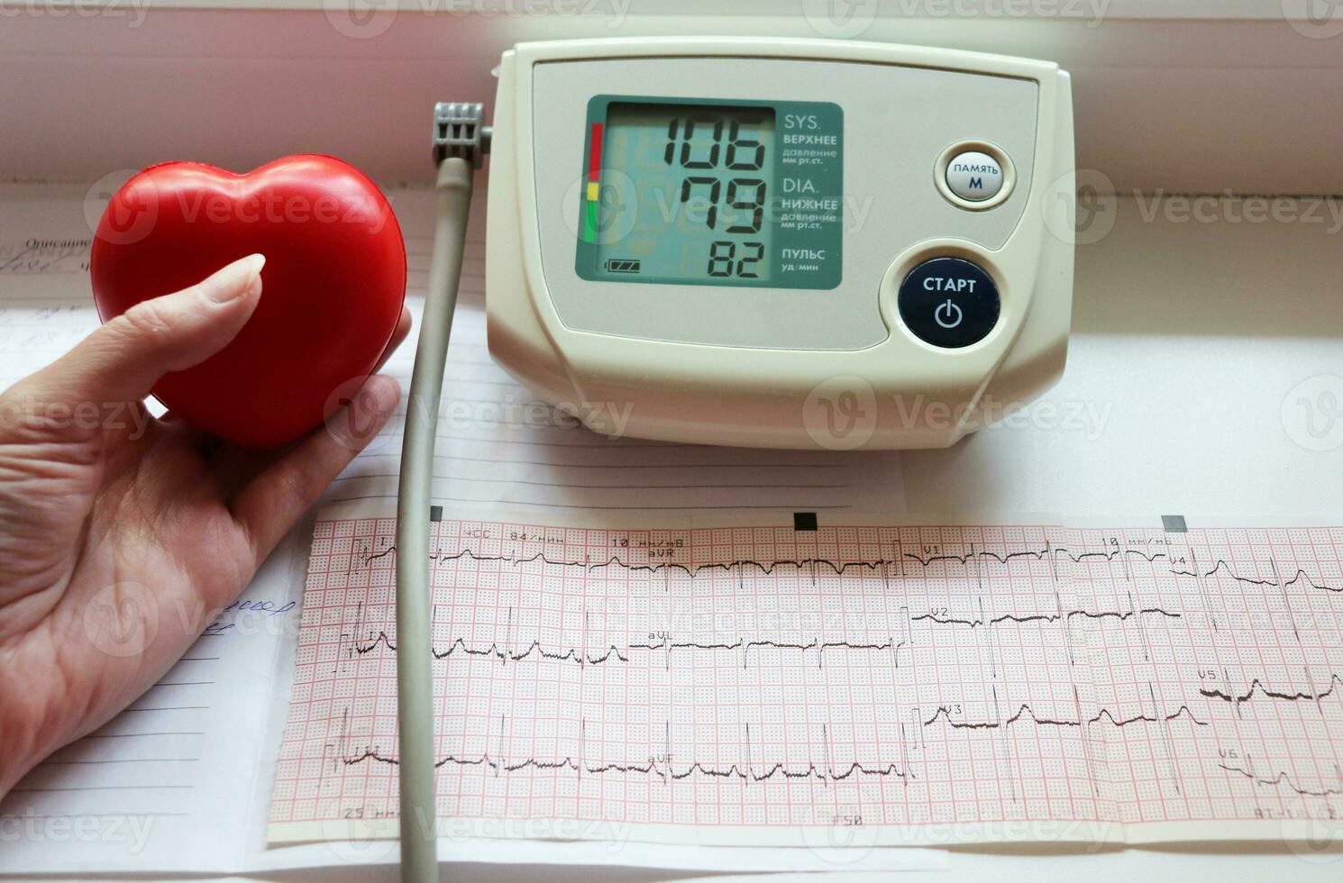 photo cardiogram results next to an electronic portable blood pressure meter - an included device with indicators on the screen and a heart-shaped rubber hand trainer in a female hand