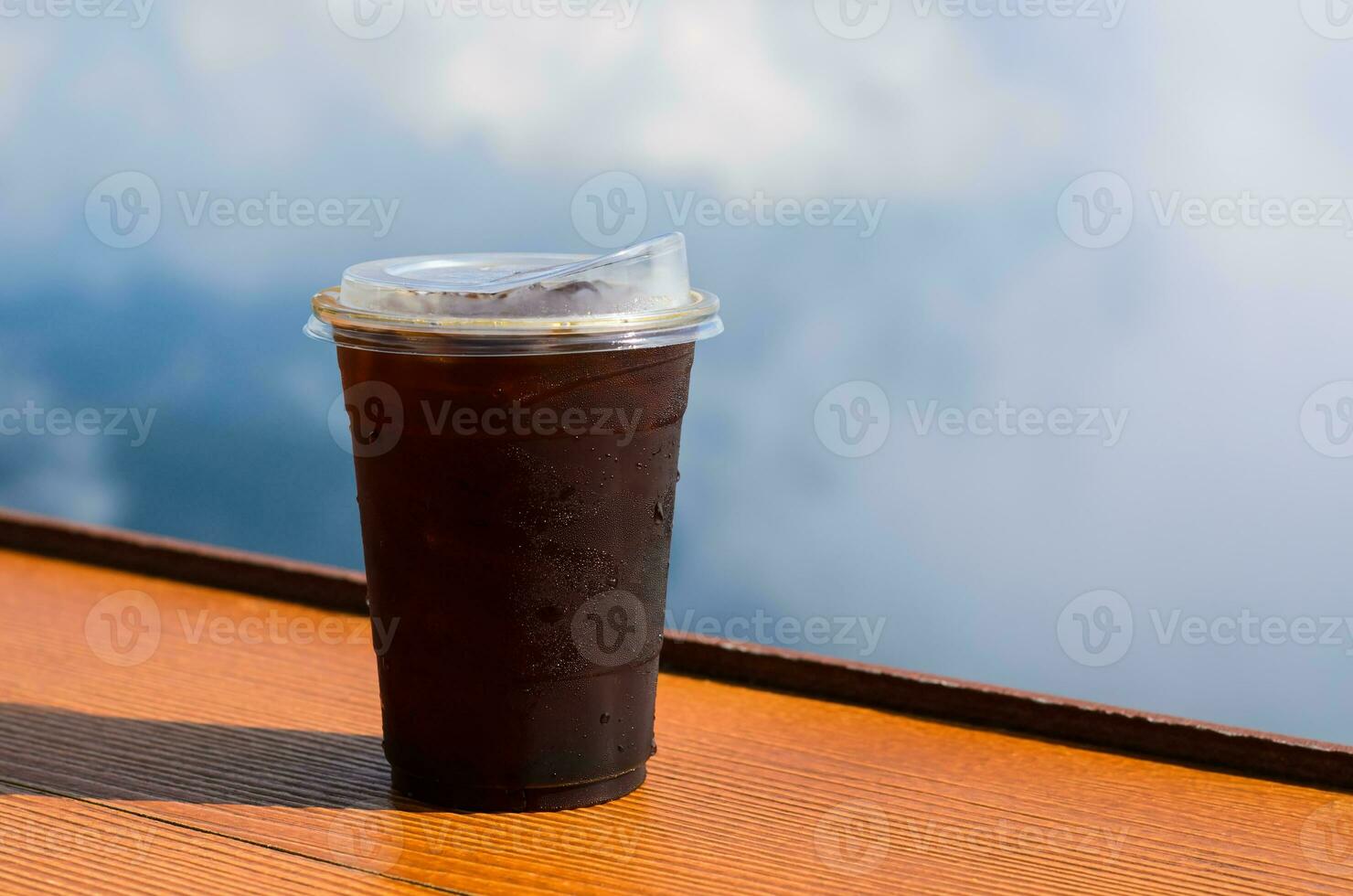 A glass of iced coffee with iced cube puts on table with sky and cloud