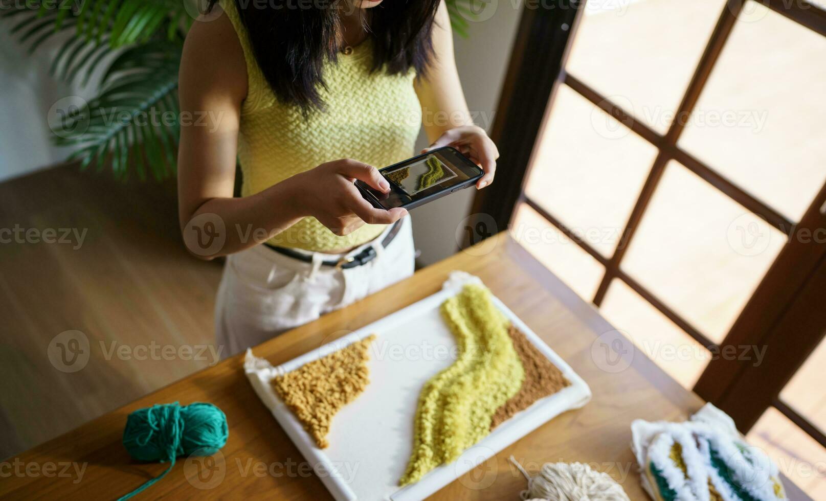 Asian Woman taking smartphone photo Punch needle. phone posting on social networks in studio workshop. designer workplace Handmade craft project DIY embroidery