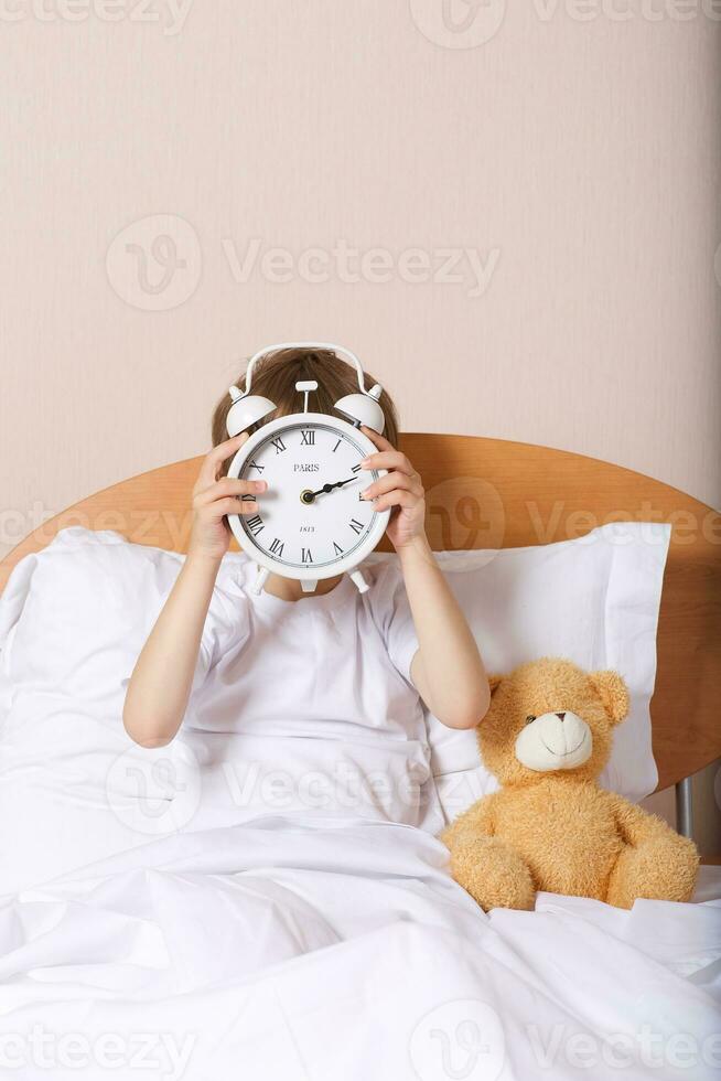 Boy is sitting in the bed with big alarm clock photo