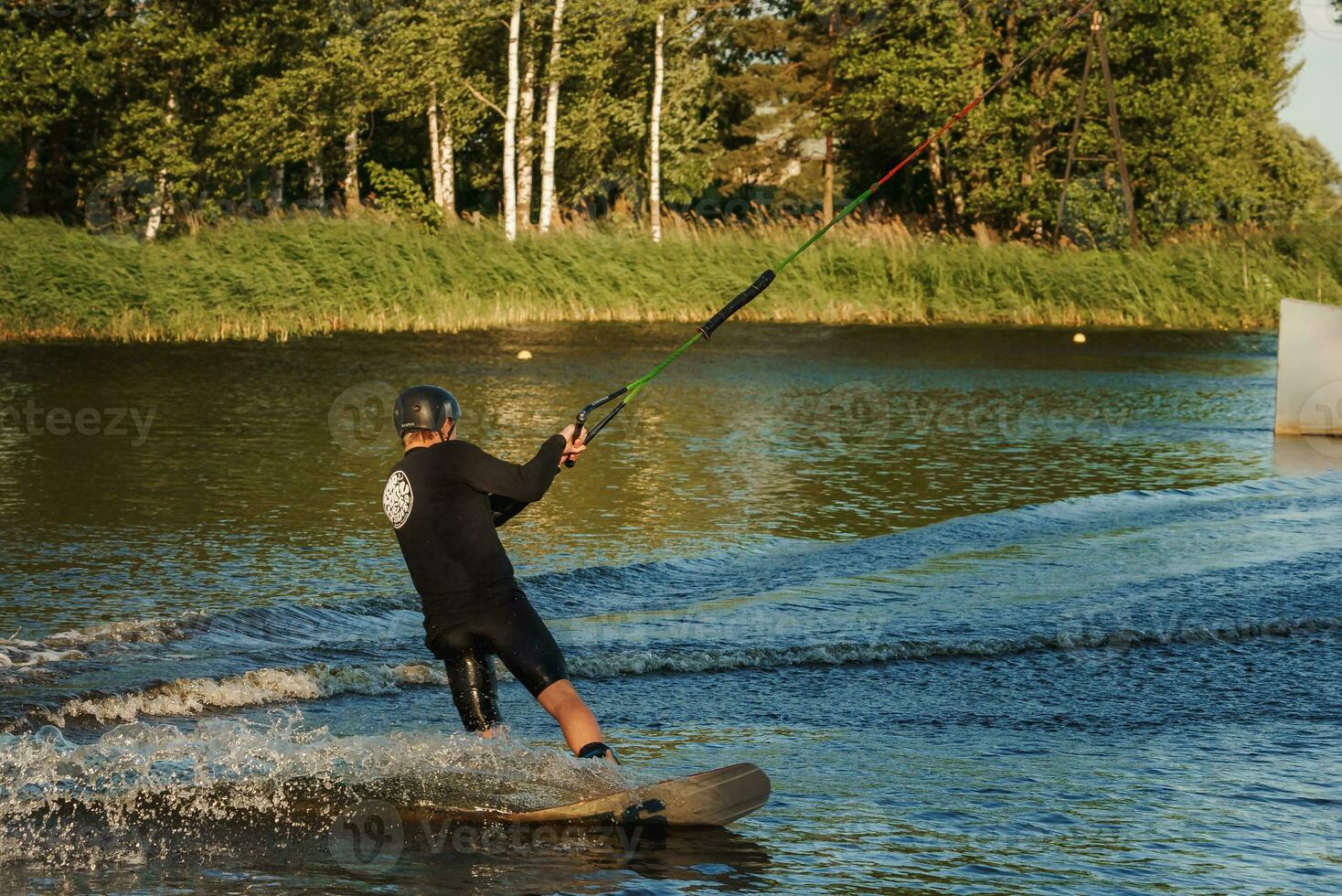 Beautiful view of young man rider holding rope and making extreme jump on wakeboard. Wakeboarding and water sports activity. Low angle shot of man wakeboarding on a lake. Man water skiing at sunset. photo