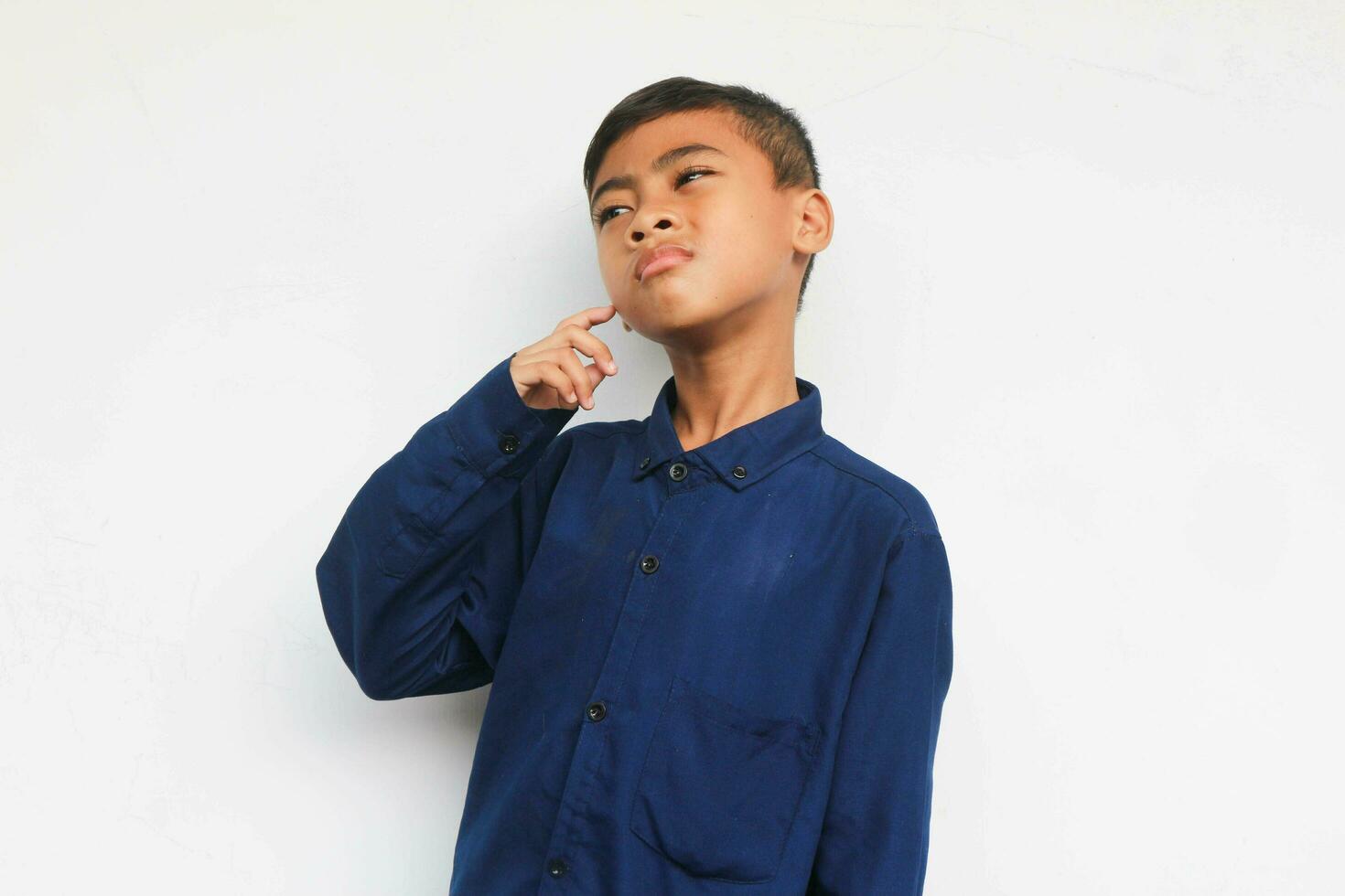 Smart kid in blue shirt touching cheek and looking up while thinking against white background photo