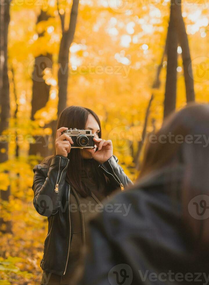 Mother taking picture her charismatic daughter on retro camera in autumn park. Hobbies and leisure concept. photo