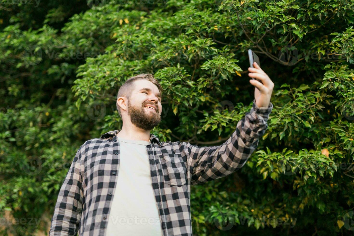 Man taking selfie portrait over tree background - Happy millennial guy enjoying summer holidays in city - Youth and technologies photo