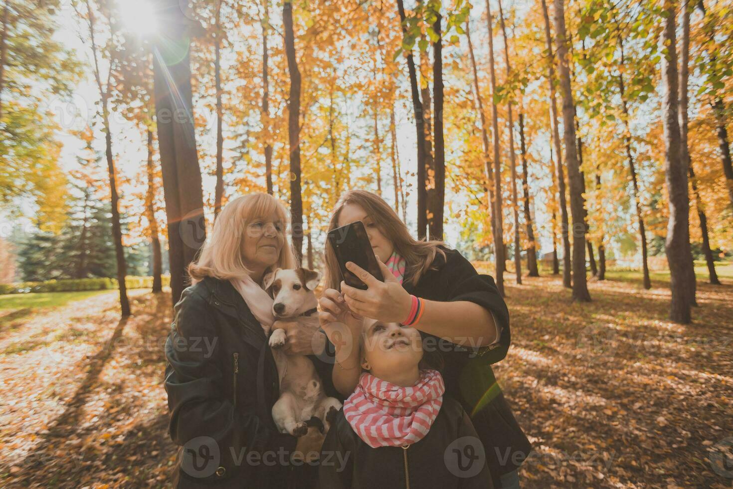Mother, grandmother and little granddaughter with jack russell terrier dog taking selfie by smartphone outdoors in autumn nature. Family, pets and generation concept photo