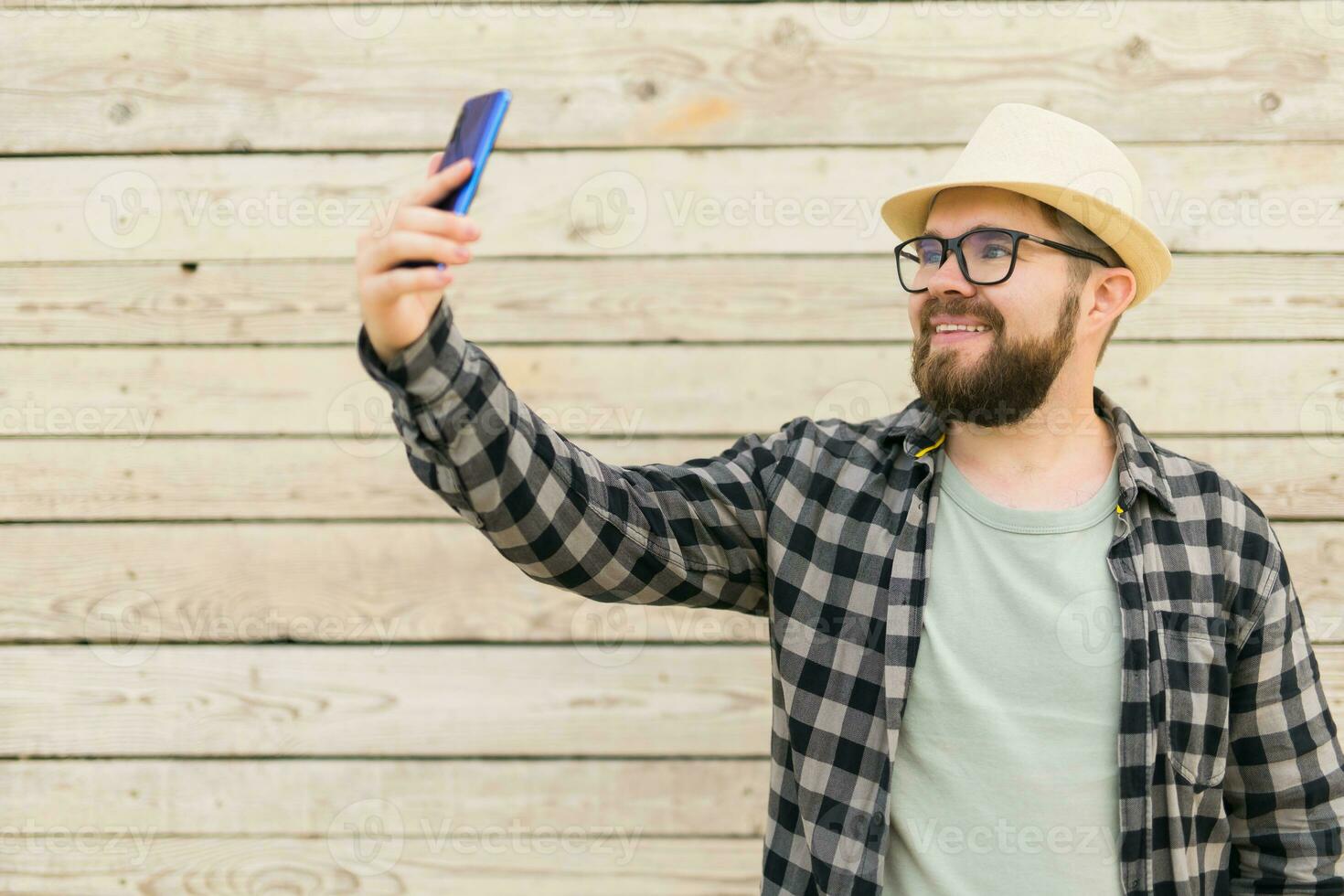Bearded man takes selfie with smartphone during summer city break over wooden wall - selfie photos and social media app concept