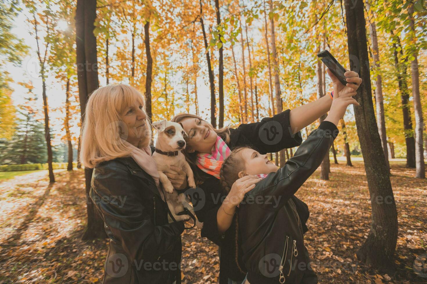 Mother, grandmother and little granddaughter with jack russell terrier dog taking selfie by smartphone outdoors in autumn nature. Family, pets and generation concept photo