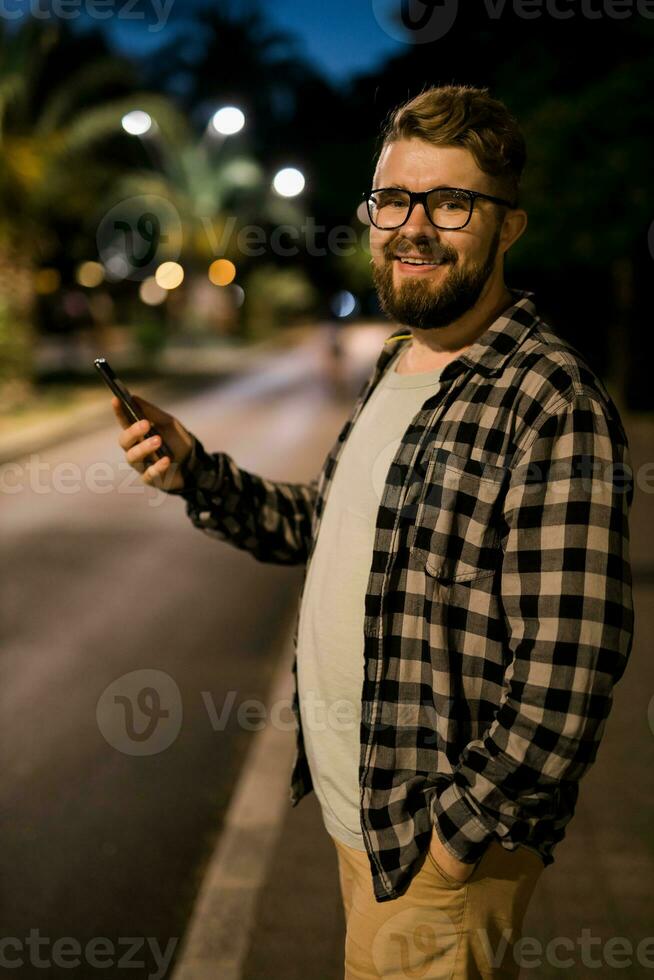 barbado hombre vistiendo los anteojos es participación desplazamiento mensajes de texto en su Teléfono móvil a noche calle. chico llamadas para Taxi en un aplicación en noche ciudad - Copiar espacio y sitio para publicidad foto