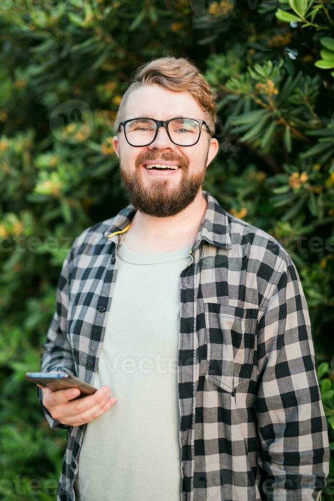 retrato de atractivo alegre chico utilizando teléfono inteligente para desplazamiento en social medios de comunicación terminado verde árbol antecedentes foto