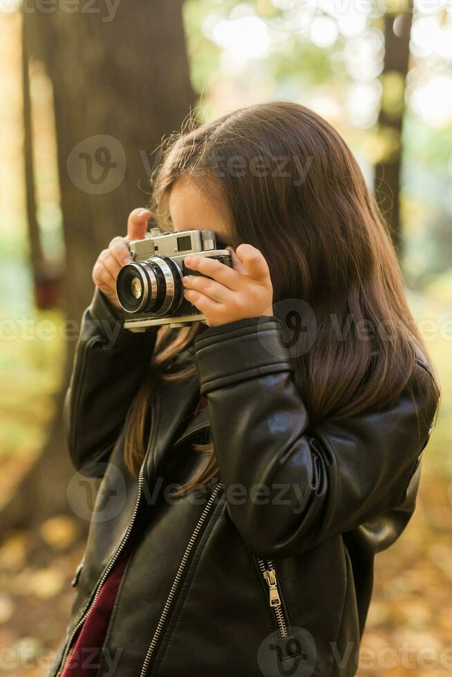 un pequeño niña tomar un foto con antiguo retro cámara en otoño naturaleza. ocio y pasatiempo concepto.