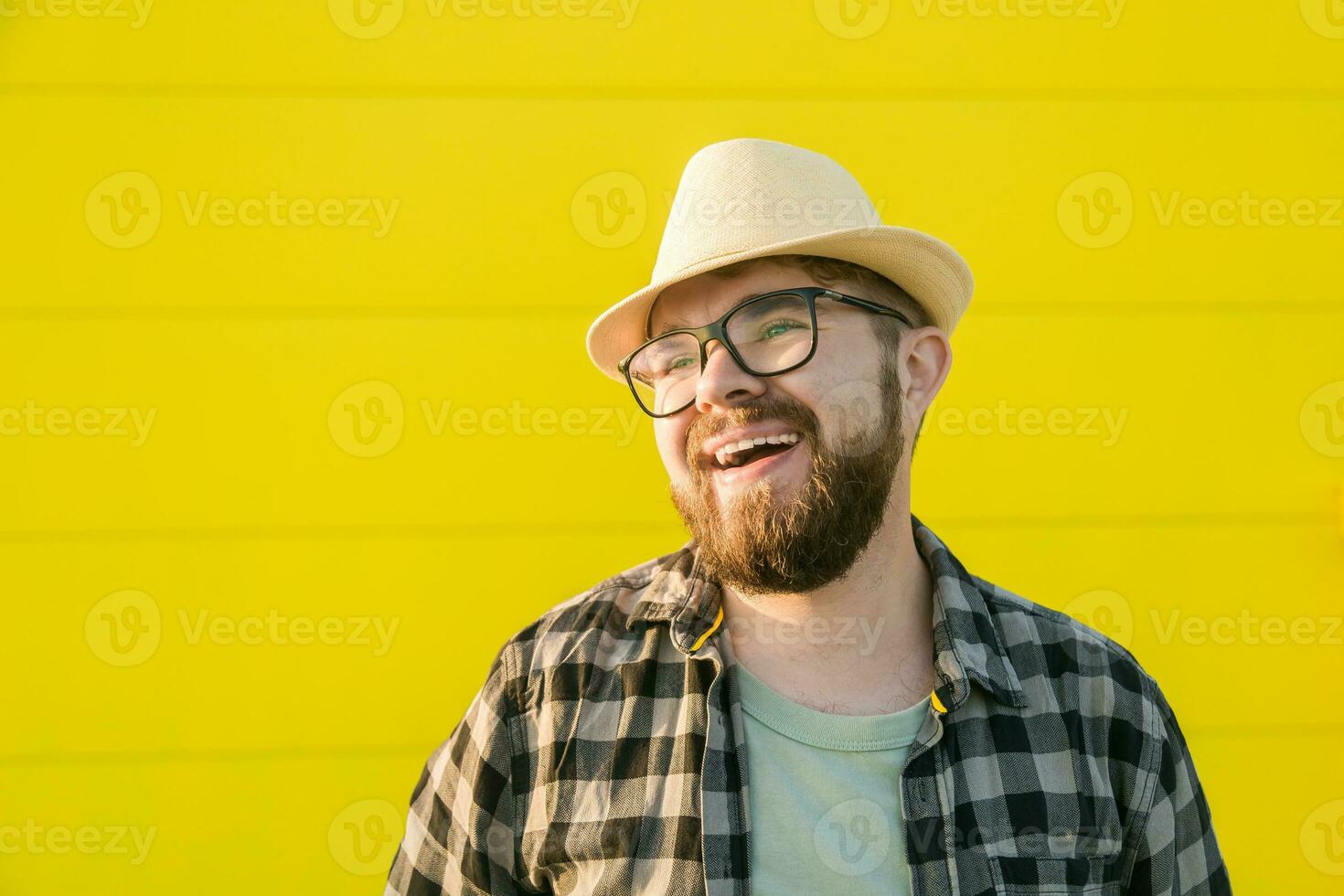 Portrait of laughing young hipster man smiling over yellow wall store outdoor background - Handsome trendy bearded guy with hat and glasses standing outdoors - Emotions and trendy youth people concept photo