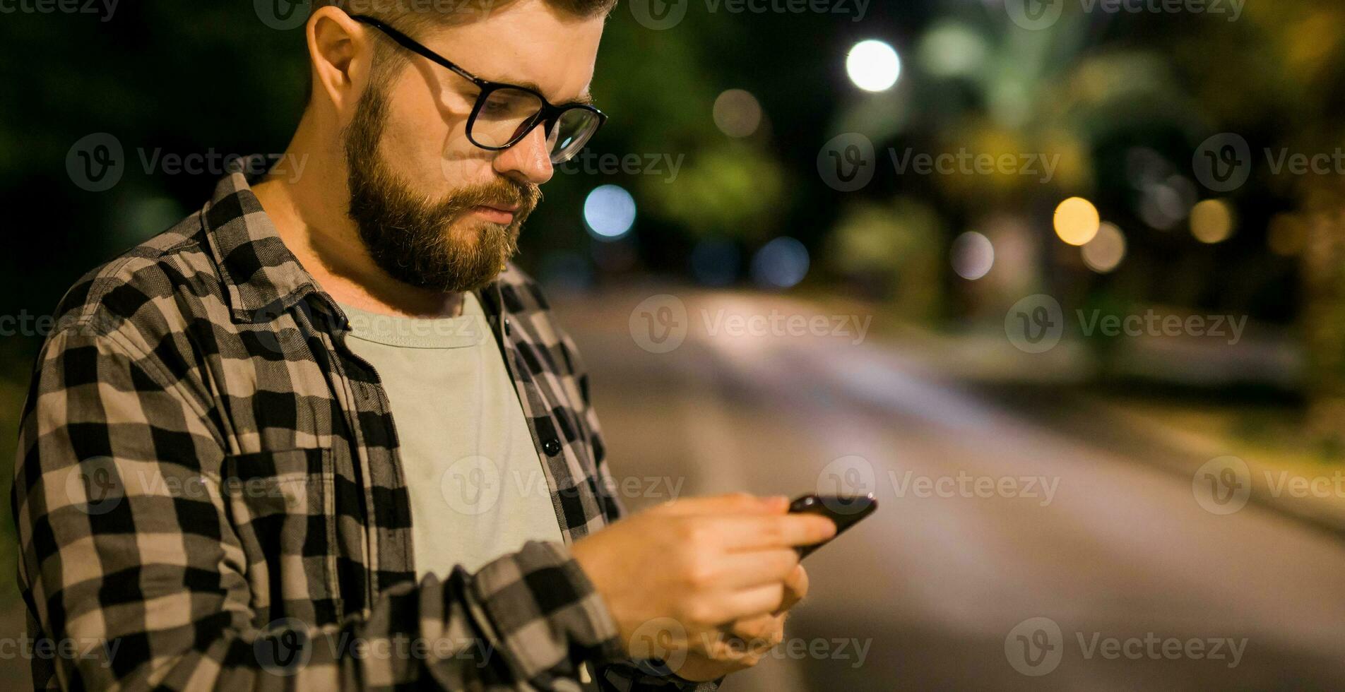 bandera barbado hombre vistiendo los anteojos es participación desplazamiento mensajes de texto en su Teléfono móvil a noche calle. chico llamadas para Taxi en un aplicación en noche ciudad - Copiar espacio y sitio para publicidad foto