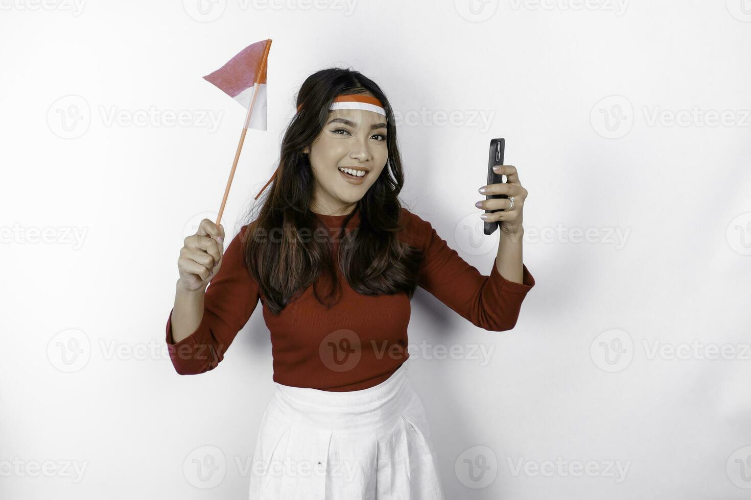 A happy Asian woman wearing headband, holding her phone and Indonesian flag, isolated by white background. Indonesia's independence day photo