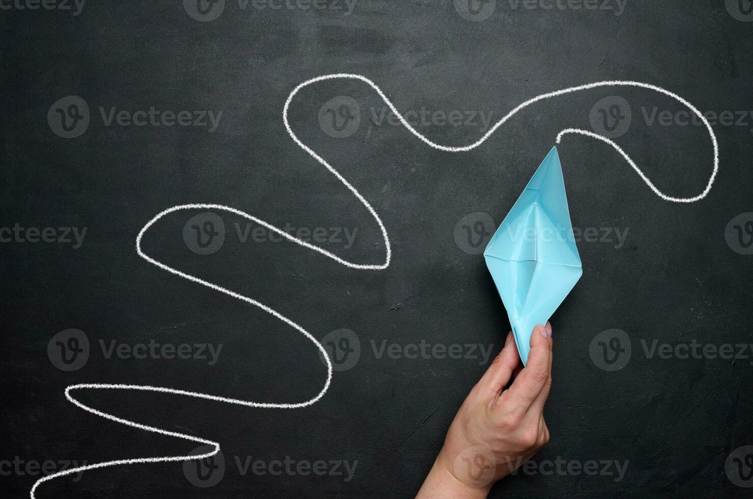 A female hand holds a paper boat against a black chalkboard background with a long path. Concept of mentorship and assistance photo