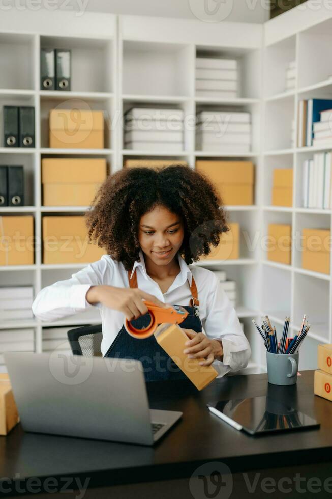 Startup small business SME, Entrepreneur owner African woman using smartphone or tablet taking receive and checking online purchase shopping order to preparing pack product box. photo