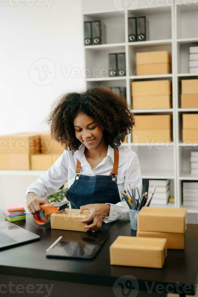 Startup small business SME, Entrepreneur owner African woman using smartphone or tablet taking receive and checking online purchase shopping order to preparing pack product box. photo