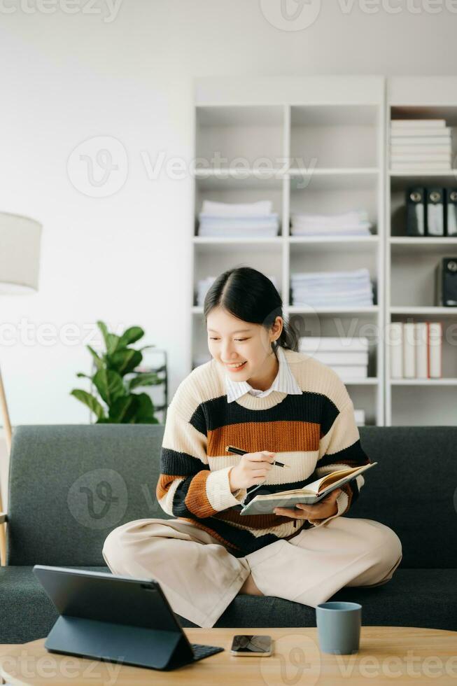 atractivo contento joven asiático estudiante estudiando a el Universidad biblioteca, sentado a sofá, utilizando un ordenador portátil computadora, tableta y auriculares teniendo un vídeo charlar. foto