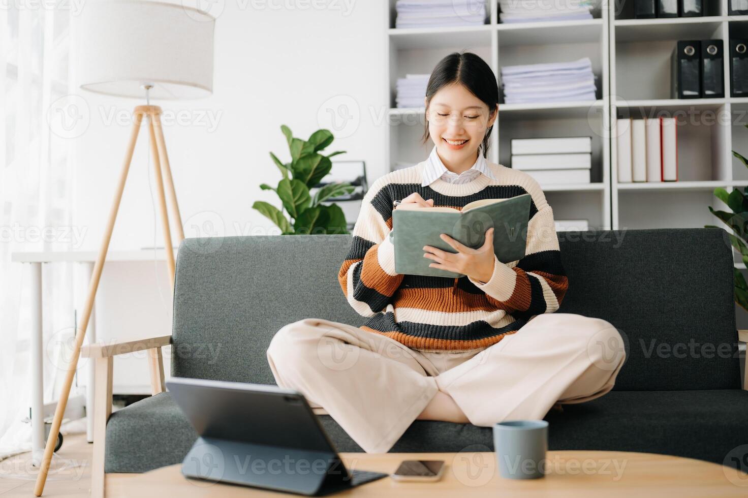 Confident Asian woman with a smile standing holding notepad and tablet in living room on the sofa at home photo
