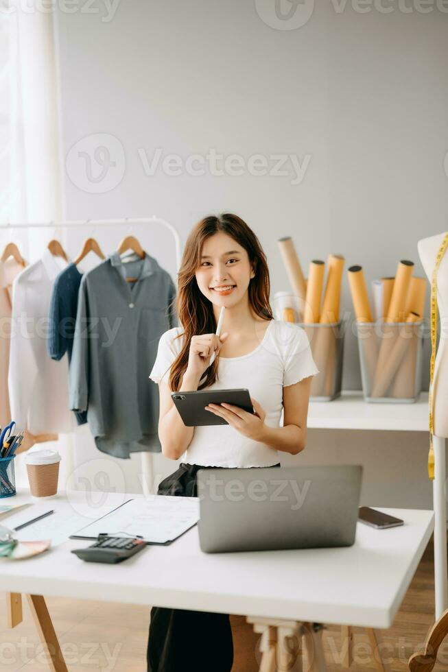 asiático Sastre mujer trabajando en ropa en sastrería taller. hermosa joven hembra Moda diseñador sonrisa y después éxito en moderno estudio foto