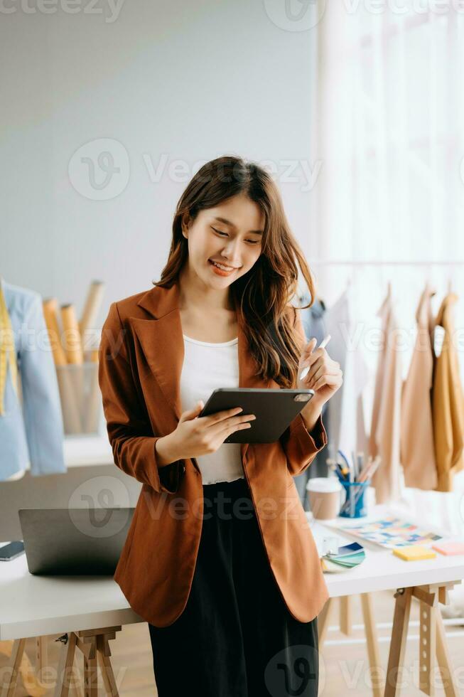 Fashion designer woman talking smart phone and using laptop with digital tablet computer in modern studio the clothes hanging on the racks morning light photo