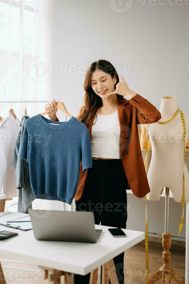 asiático joven mujer en escritorio en oficina de Moda diseñador y sostiene tableta, ordenador portátil y teléfono inteligente en blanco mesa foto