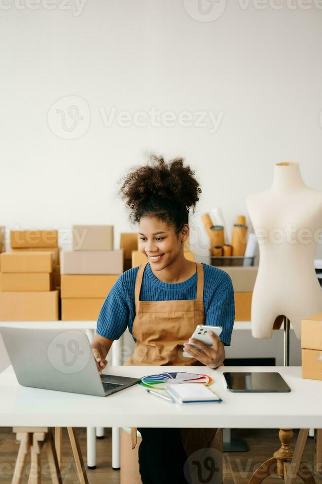Startup small business SME, Entrepreneur owner African woman using smartphone or tablet taking receive and checking online purchase shopping order to preparing pack product box. in office photo