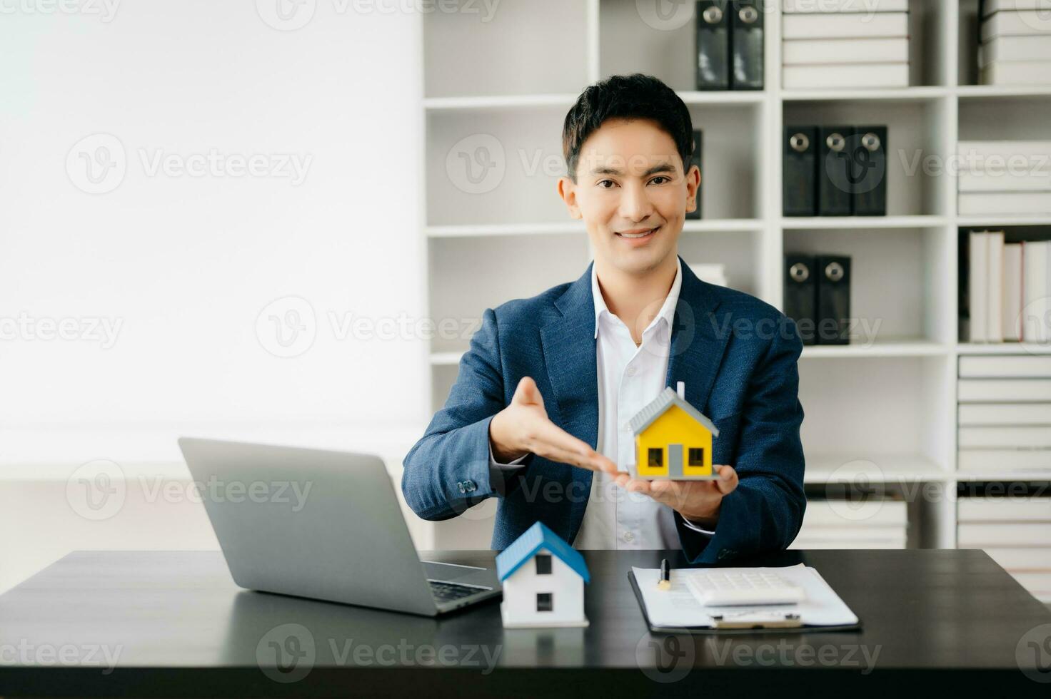 joven real inmuebles agente trabajador trabajando con ordenador portátil y tableta a mesa en moderno oficina y pequeño casa junto a él. foto