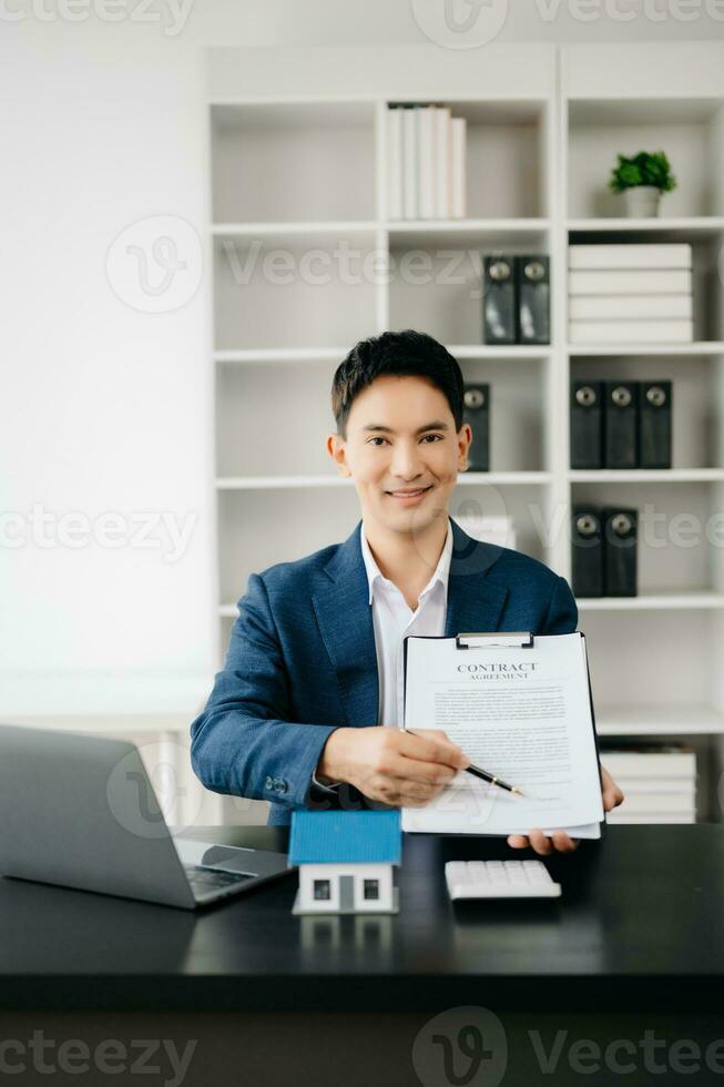 Young real estate agent worker working with laptop and tablet at table in modern office and small house beside it. photo