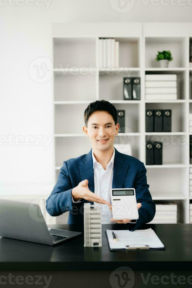 Young real estate agent worker working with laptop and tablet at table in modern office and small house beside it. photo