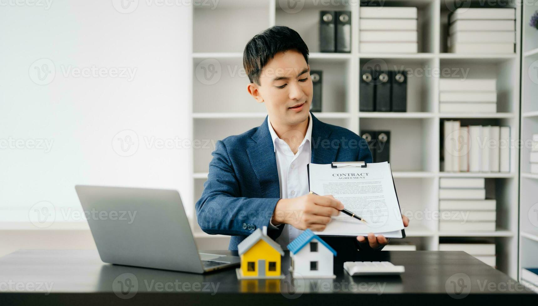 Young real estate agent worker working with laptop and tablet at table in modern office and small house beside it. photo