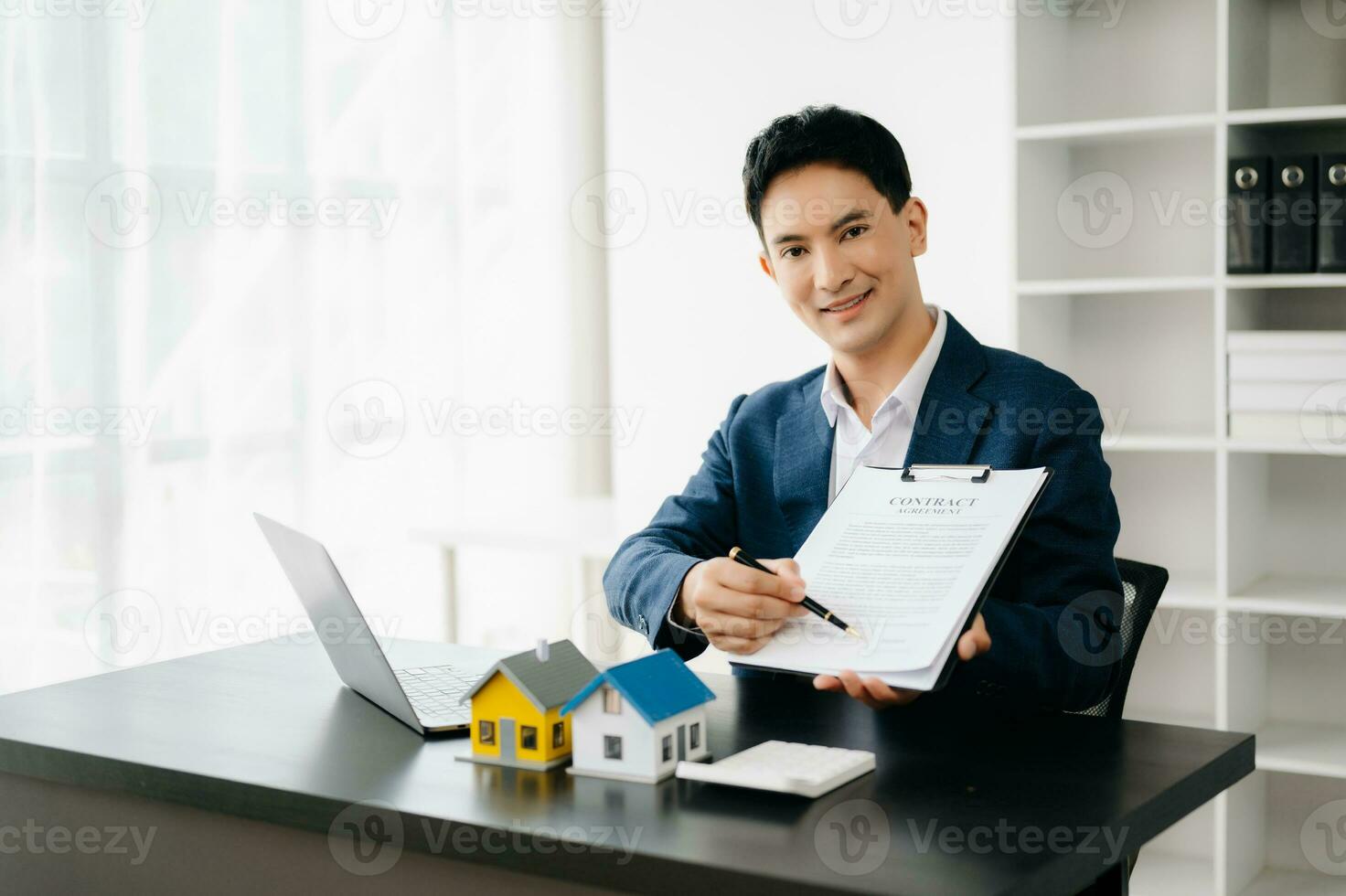 Young real estate agent worker working with laptop and tablet at table in modern office and small house beside it. photo