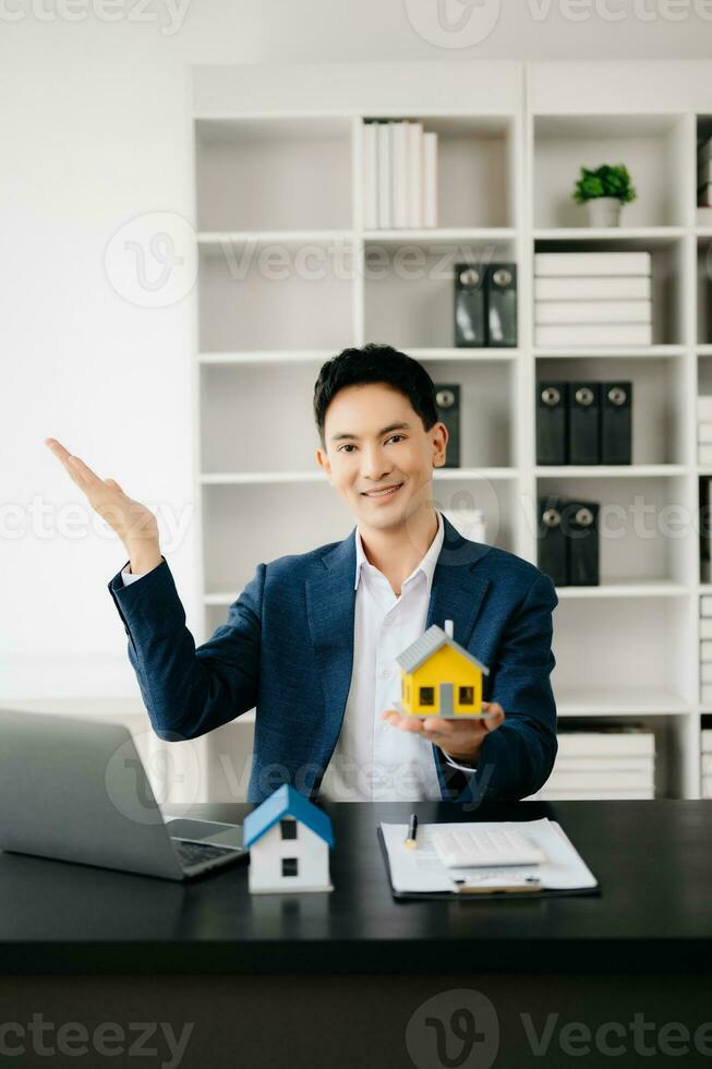 Young real estate agent worker working with laptop and tablet at table in modern office and small house beside it. photo