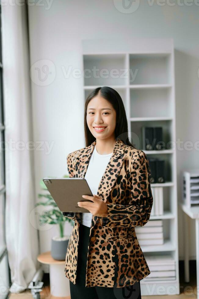 Young attractive Asian female office worker business suits smiling at camera with working notepad, tablet and laptop documents photo