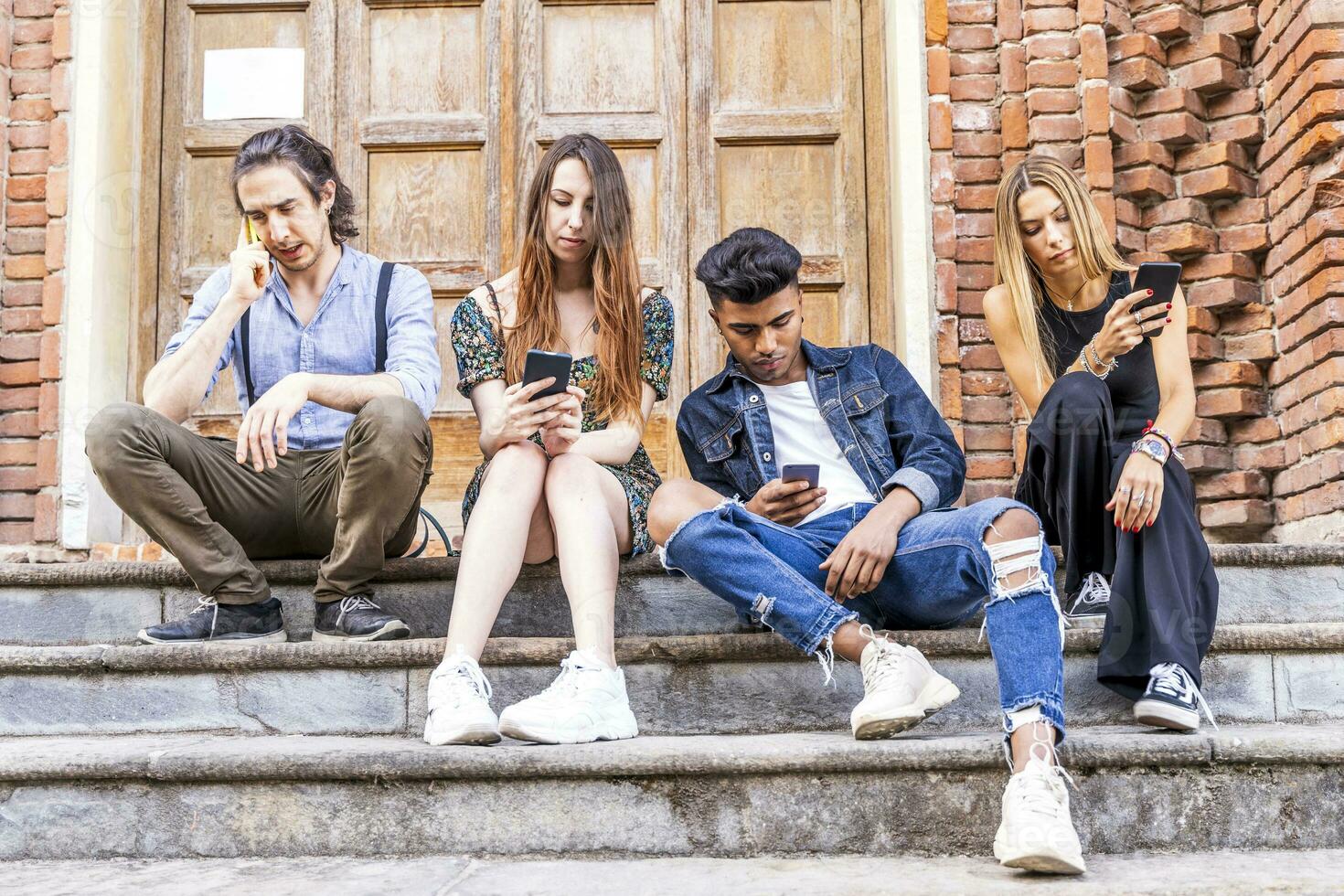 group of multiracial friends sitting on a marble staircase using smartphones photo